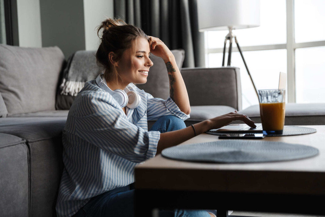 Jung woman in her living room, sitting on the floor in front of a coffee table, drinking juice and using her laptop